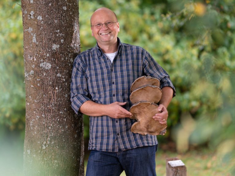 Volker Walther, Leiter des Pilzkundemuseums fuehrt Gruppen durch das Museum und hilft Gaesten bei der Bestimmung von Pilzen. Er lehnt mit einem Pilz unter dem Arm an einem Baum.